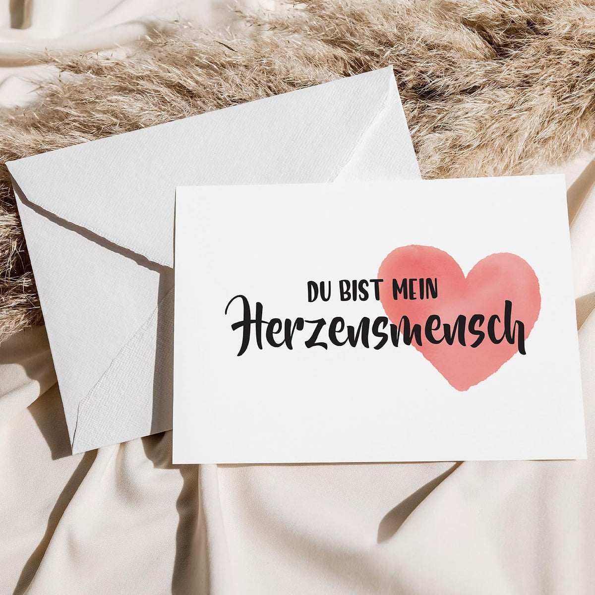 Weiße Grußkarte mit der Aufschrift „Du bist mein Herzensmensch“ in schwarzer Schrift und einem zartrosa Aquarell-Herz, die gegen einen passenden weißen Umschlag auf cremefarbenem Fabric mit getrocknetem Pampasgras gelehnt ist.