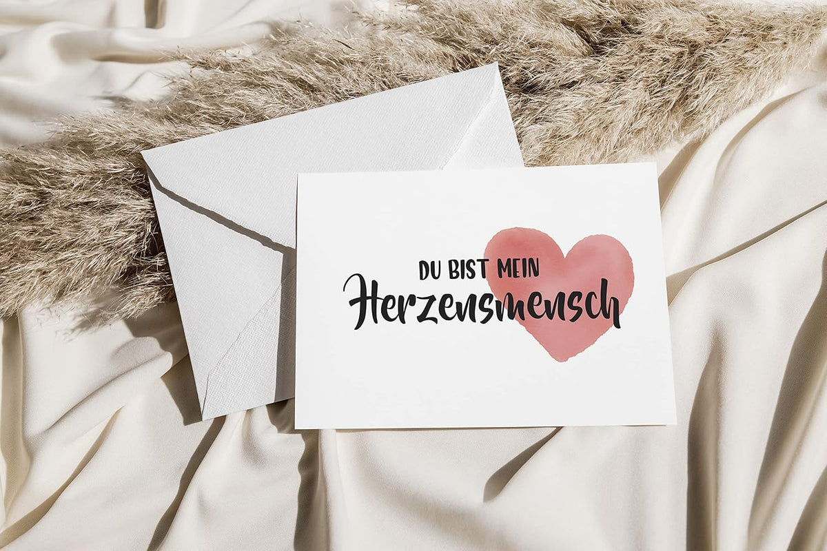 Weiße Grußkarte mit der Aufschrift „Du bist mein Herzensmensch“ in schwarzer Schrift und einem zarten rosafarbenen Aquarell‑Herz, liegt auf einem passenden weißen Umschlag oben auf cremefarbenem Fabric mit getrocknetem Pampasgras.